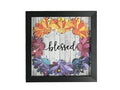 Framed decorative wall art with layered paper flowers in orange, yellow, pink, purple, and blue around the word 'blessed' on a whitewashed wood background, shown in a black frame against a gray textured wall.