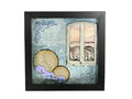 A square black-framed mixed-media art piece featuring a distressed blue background, a white window with pink sky, two gold circles, and purple flowers, displayed on a textured gray surface.